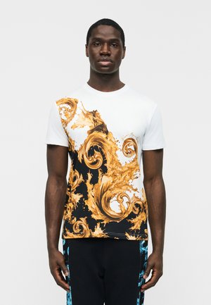 Man draagt een wit T-shirt met korte mouwen en gouden decoratieve krulpatronen op een zwarte achtergrond, staand tegen een eenvoudige lichte achtergrond.