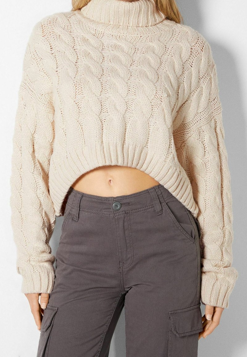 Femme portant un pull court à col roulé crème en maille torsadée et un pantalon cargo gris foncé, montrant le ventre et adoptant une posture détendue.