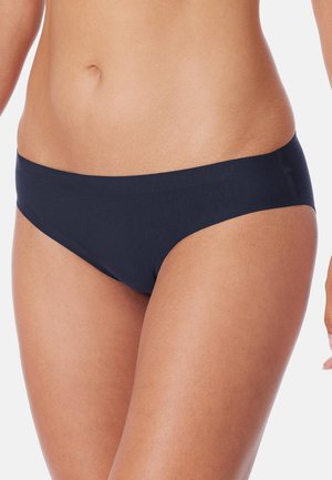 Torace femminile che indossa slip bikini senza cuciture blu scuro a vita bassa su sfondo bianco semplice.