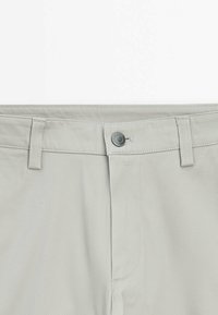 Lichtgrijze broek met een gladde textuur, sluiting met voorknop en riemlussen. Bevat een subtiel visgraatpatroon in de stof.