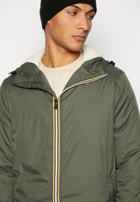 Green lightweight jacket with a zip closure, featuring a white fleece-lined hood, and colored stripes along the zipper.