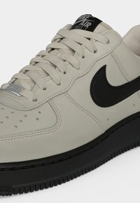 Μπεζ αθλητικό παπούτσι Nike Air Force 1 με μαύρο λογότυπο swoosh, μαύρη σόλα, μπεζ κορδόνια και διάτρητη περιοχή στα δάχτυλα.