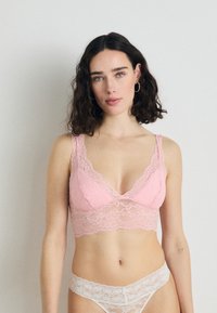 Krajkový bralet v svetloružovej farbe so širokými ramienkami a vykrojenými okrajmi, doplnený o zodpovedajúce krajkové nohavičky, ktoré sa vyznačujú jemnou textúrou a delikátnym vzorom.