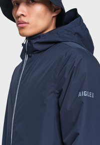 Jeune homme portant une veste imperméable bleu marine avec capuche et un chapeau bob assorti, montrant partiellement le visage et le haut du corps.