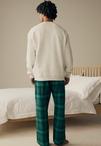 Harmaa huppari ja vihreät ruudulliset housut, rento malli, pehmeä kangas, pyöreä kaula-aukko ja pitkät hihat. Taustalla on sänky valkoisine vuodevaatteineen.