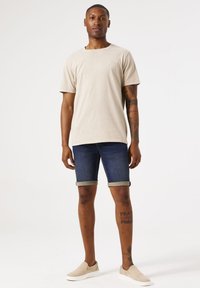 Camiseta de algodón beige de mangas cortas, combinada con pantalones cortos de mezclilla azul oscuro con dobladillos enrollados, y zapatillas beige claras sin cordones.