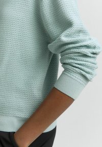 Šviesiai mėtų žalios spalvos megztinis su tekstūruotu, korio rašto audiniu. Suapvalinti rankovės kraštai ir banguota apačia, derinamas su tamsia medžiaga po apačia.