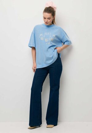 Vrouw draagt een oversized lichtblauw T-shirt met bloemenprint en donkerblauwe wijde spijkerbroek, staand tegen een witte muur.