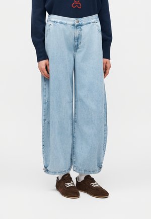 Άτομο που φοράει ανοιχτό μπλε τζιν παντελόνι με φαρδιά μπατζάκια σε κοντή γραμμή, σκούρα καφέ αθλητικά παπούτσια με λευκά κορδόνια, λευκές κάλτσες και μπλε σκούφο με κόκκινη κεντητική.