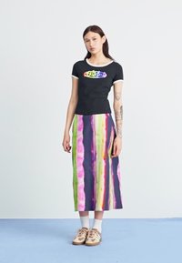 Sort kortærmet t-shirt med regnbue-logo, parret med et langt, farverigt stribede nederdel i pink, grøn og lilla. Brune sneakers båret.