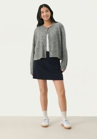 Cardigan en tricot gris avec des boutons, porté sur un haut blanc et une jupe marine. Caractérisé par une coupe décontractée, des manches longues et des baskets blanches avec des semelles beige.