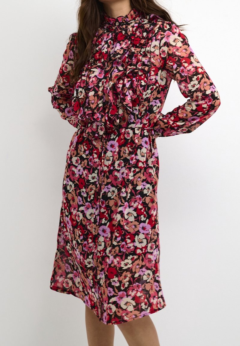 Kukallinen midi-mekko, jossa on frillakaulus, pitkät hihat ja kapeampi vyötärö. Kangas on kevyt, ja siinä on vaaleanpunaisia, punaisia ja beigejä kukkia mustalla taustalla.