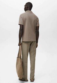 Camisa polo beige de manga corta, combinada con pantalones caqui claros. Sosteniendo una bolsa de lona beige, de pie sobre un fondo liso.