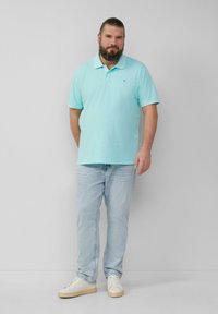 Lichtblauw polo-shirt met korte mouwen, kraag, gedragen met lichte denim jeans en witte sneakers, staande tegen een grijze achtergrond.