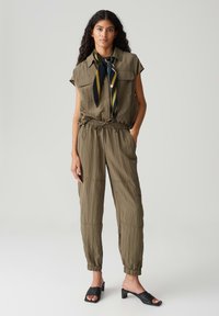 Olijfgroene jumpsuit met korte mouwen, strikceintuur en taps toelopende benen. Gecombineerd met een blauw en geel patroon sjaal en zwarte sleehakken.