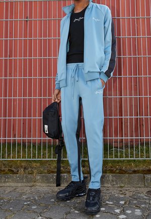 Personne portant un survêtement bleu clair avec des bandes noires, des baskets noires et tenant un petit sac noir, debout près d'une clôture métallique.