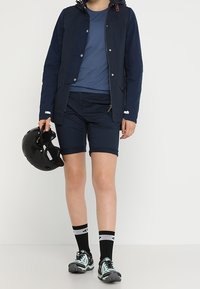 Personne portant une veste navy, une chemise bleue, un short navy, des chaussettes noires et des baskets, tenant un casque de vélo noir dans une main.