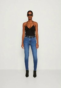 Top halter preto com decote em V, combinado com jeans skinny azuis e botas de tornozelo pretas. Modelo usa óculos de sol oversized pretos.