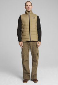Gilet imbottito color khaki con colletto alto, dotato di zip frontale e logo del marchio, abbinato a pantaloni cargo marroni e sneakers.