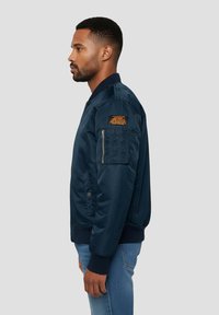 Veste bomber marine avec une poche zippée sur le bras, des poignets côtelés et un patch textile. Texture lisse, design classique et silhouette ajustée.