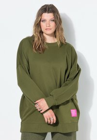 Sweatshirt oversize vert olive avec des manches longues, des poignets côtelés et un ourlet texturé. Présente une étiquette rose avec du texte blanc sur le côté.