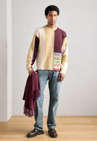 Kootud džemper plaastrilise mustriga beezhibi, bordoo ja kollase toonides. Kaasas bordoopunane sall ja sinised teksapüksid. Pruunid kingad.