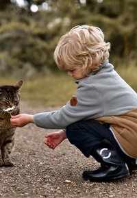 Child in gray fleece jacket and brown accents kneels on a gravel path, offering a white flower to a tabby cat. Black rubber boots are visible.
