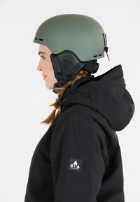 Matte olijfgroene helm met ventilatiegaten en zwarte oorkussens, gedragen met een zwarte jas met een logo op de mouw.