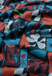 Tkanina z abstraktnimi geometrskimi in cvetličnimi vzorci v modri, rdeči, črni, beli in zeleni barvi, ki prikazuje različne oblike in plastne teksture.