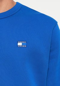 Sininen collegepaita, joka on valmistettu strukturoituneesta kankaasta; siinä on ribbed-kaulaosa ja pieni suorakulmainen logopatch, jossa lukee "TOMMY JEANS" edessä.