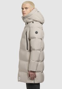 Chaqueta puffer beige con capucha ajustable, cremallera frontal y bolsillos, textura acolchada y parche con logotipo en la manga.