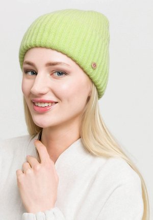 Zöld bordázott kötött sapka texturált felülettel és egy kis fém logóval. A modellen egy világos pulóver van, enyhén mosolyog.