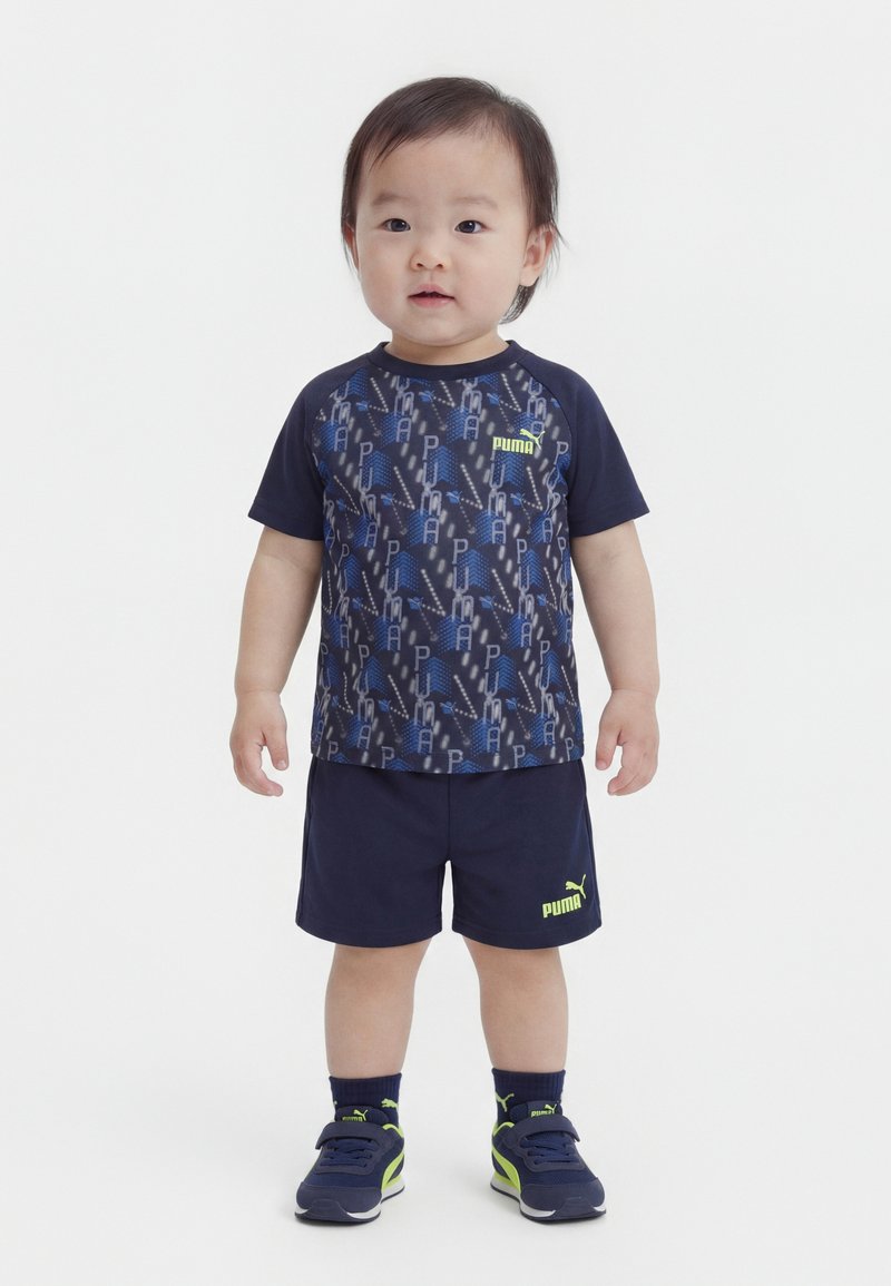 Νήπιο όρθιο, φοράει μπλε σκούρο μπλουζάκι Puma με μπλε σχέδιο, αντίστοιχα σορτς, μπλε σκούρες κάλτσες και μπλε σκούρα αθλητικά παπούτσια με νέον λεπτομέρειες.