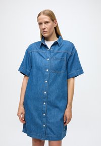 Rifľové šaty-košeľa s krátkymi rukávmi, gombíkmi vpredu, náprsným vreckom a voľným strihom. Vyrobené z modrej bavlnenej látky s jemnou textúrou.