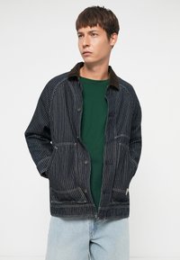 Striped denim jacket with brown corduroy collar, button closure, and front pockets, worn over a green shirt and blue jeans.