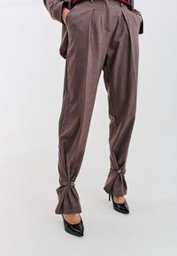 Hnedé nohavice na mieru so zapínaním na gombíky, zúžené na členkoch, nosené s čiernymi lodičkami s ihličkovým podpätkom a hnedým zladeným sakom.