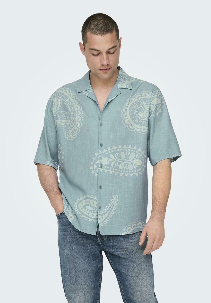 Kurzärmliges Hemd in hellblauem Stoff mit weißen Paisleymustern; besitzt einen Kragen und eine lockere Passform. Kombiniert mit blauen Jeans.