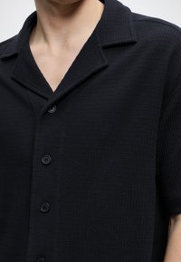 Chemise noire texturée à manches courtes avec col cranté, portée par une personne montrant le cou et le haut du torse.