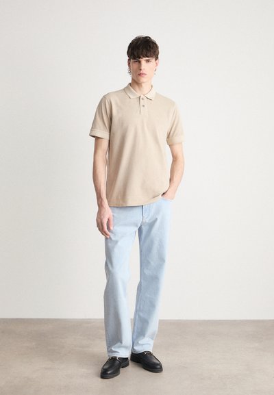 Polo shirt en coton beige avec un col et trois boutons, associé à un jean droit bleu clair et des chaussures noires. Design simple, texture douce.