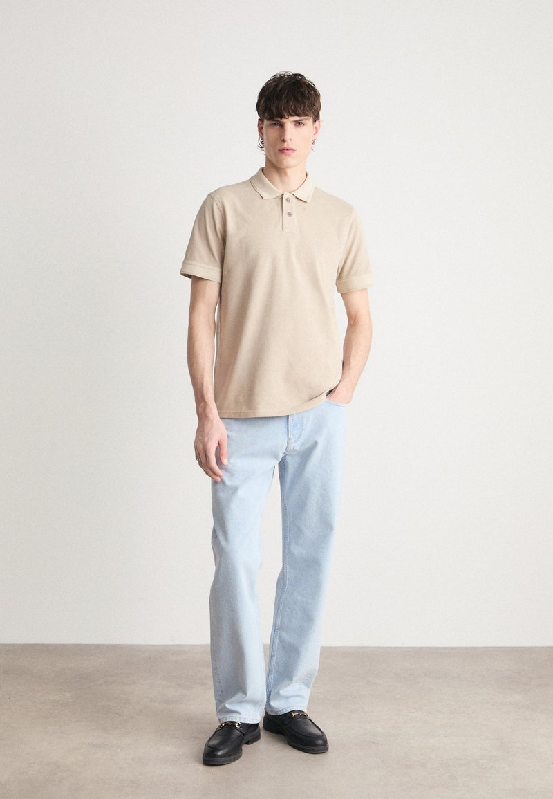 Camisa polo de algodón beige con collar y tres botones, combinada con jeans de pierna recta de color azul claro y zapatos negros. Diseño sencillo, textura suave.