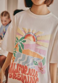Vit T-shirt med en färgglad grafik av en sol, palmer, vågor och texten "California Dreaming" i fet, lekfull typografi.