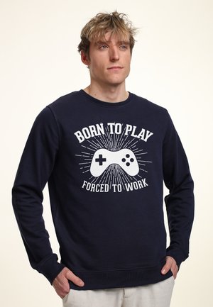 Marineblå sweatshirt med et grafisk design af en spilcontroller med hvid tekst "BORN TO PLAY FORCED TO WORK," rund hals, lange ærmer.