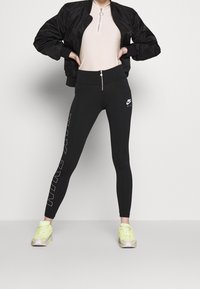 Person som bär svarta Nike Air leggings med vita logotyper, neongula sneakers, en beige topp och en svart bomberjacka, stående med händerna på höfterna.