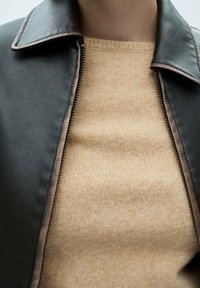 Schwarze Lederjacke mit Kragen und seitlichem Reißverschluss, über einem beigen Strickpullover mit weicher Textur geschichtet.