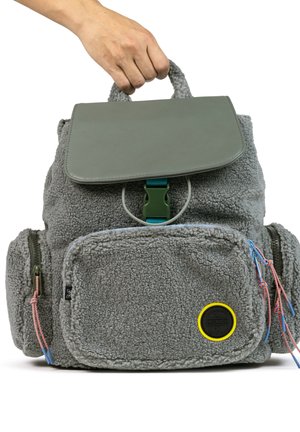 Mochila de felpa gris con una solapa de cuero verde, detalles azules, bolsillo frontal y un asa. Incluye cremalleras y un parche de logo amarillo.