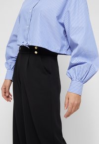 Cropped blue and white striped shirt with long puff sleeves, tie detail at the hem, paired with high-waisted black trousers with gold buttons.