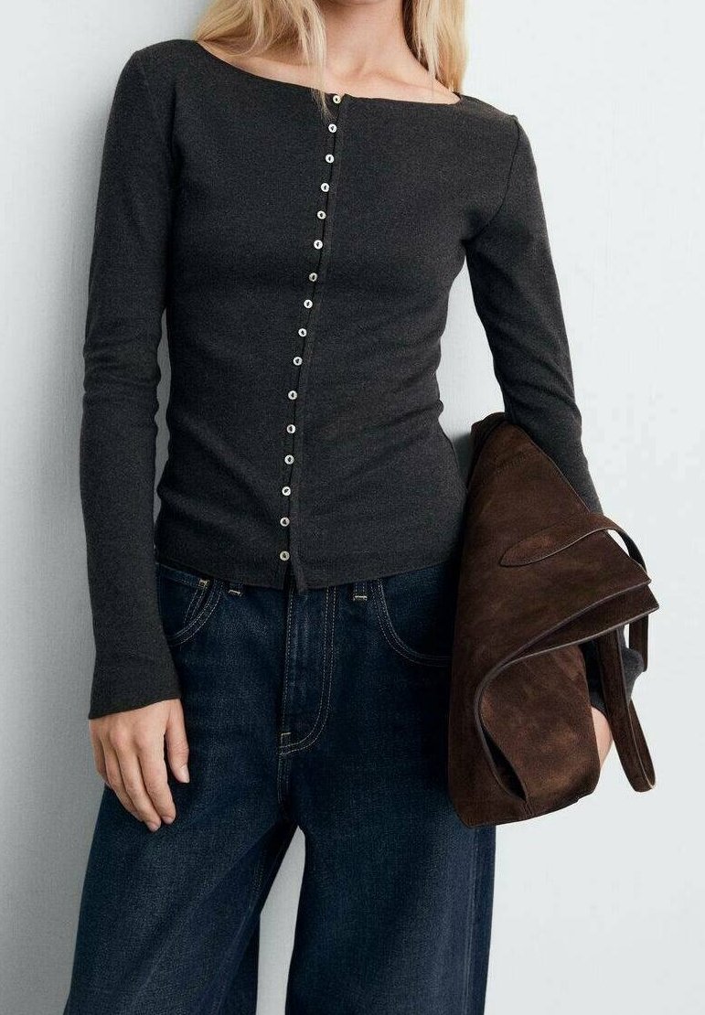 Femme portant un haut ajusté gris foncé à manches longues boutonné et un jean large foncé, tenant un grand sac plié en daim marron sous le bras.