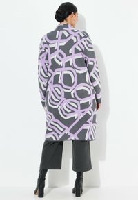 Langer, grauer Cardigan mit geometrischem Muster in Lila und Weiß, mit tailliertem Kragen und seitlichen Schlitzen. Getragen über grauen Hosen und schwarzen Stiefeln.