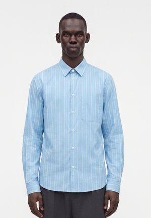 Homme portant une chemise bleu clair à manches longues avec des rayures verticales blanches, boutonnée, debout devant un fond blanc uni.