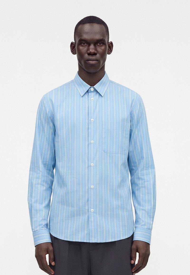 Homme portant une chemise bleu clair à manches longues avec des rayures verticales blanches, boutonnée, debout devant un fond blanc uni.
